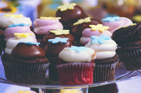 A group of decorative cupcakes, each topped with colorful frosting and floral-shaped sprinkles. The cupcakes are arranged on a glass stand, displaying a variety of chocolate and vanilla cakes with pink, white, and brown frosting.