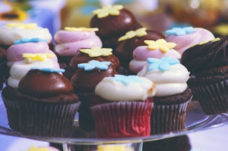 A group of decorative cupcakes, each topped with colorful frosting and floral-shaped sprinkles. The cupcakes are arranged on a glass stand, displaying a variety of chocolate and vanilla cakes with pink, white, and brown frosting.