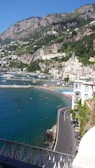 A scenic coastal view features a winding road alongside crystal-clear blue waters. On the right, a cliffside town with charming, pastel-colored buildings is nestled against a backdrop of lush green hills and rugged mountains. The beach below is dotted with vibrant blue and red umbrellas, with numerous people enjoying the sun.