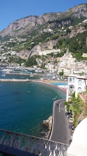 A scenic coastal view features a winding road alongside crystal-clear blue waters. On the right, a cliffside town with charming, pastel-colored buildings is nestled against a backdrop of lush green hills and rugged mountains. The beach below is dotted with vibrant blue and red umbrellas, with numerous people enjoying the sun.