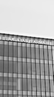 A modern building with a glass facade featuring vertical and horizontal lines creating a grid pattern. The top of the building displays the text 'FAKULTAS FARMASI' in bold, capital letters.