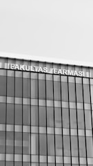 A modern building with a glass facade featuring vertical and horizontal lines creating a grid pattern. The top of the building displays the text 'FAKULTAS FARMASI' in bold, capital letters.