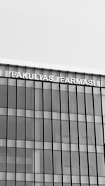 A modern building with a glass facade featuring vertical and horizontal lines creating a grid pattern. The top of the building displays the text 'FAKULTAS FARMASI' in bold, capital letters.
