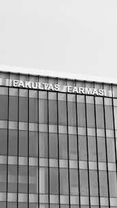 A modern building with a glass facade featuring vertical and horizontal lines creating a grid pattern. The top of the building displays the text 'FAKULTAS FARMASI' in bold, capital letters.