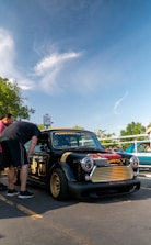 A small, customized black car with gold details and the words 'Powered by Honda' on the windshield. Two people are inspecting the car, which has a low profile and gold rims. The sky is clear with some white clouds and a tree is in the background.