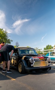 A small, customized black car with gold details and the words 'Powered by Honda' on the windshield. Two people are inspecting the car, which has a low profile and gold rims. The sky is clear with some white clouds and a tree is in the background.