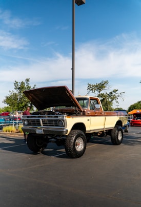 A vintage cream and brown pickup truck with its hood open is parked in an outdoor setting. The truck is lifted with large tires and is situated in a parking lot on a sunny day with a clear blue sky. There are trees and other cars in the background.