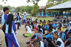 A group of young people, mostly wearing blue and black uniforms, are gathered outdoors. They are sitting and standing on a sandy area surrounded by trees. Some individuals are holding papers, suggesting a group activity or discussion. Nearby, there are blue and yellow buildings and a metal shelter with more people sitting underneath.