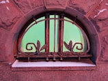 An arched window with decorative metal bars featuring scroll and leaf designs. The window is set in a textured brick wall, and the glass has a green tint, reflecting light. There is a dusting of snow on the windowsill.