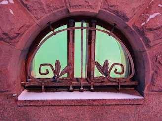 An arched window with decorative metal bars featuring scroll and leaf designs. The window is set in a textured brick wall, and the glass has a green tint, reflecting light. There is a dusting of snow on the windowsill.