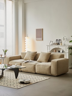 A modern living room features a large beige sofa adorned with three cushions, a coffee table with various items including a candle and vase, and a beige rug with a dotted pattern. A robotic vacuum cleaner is on the floor. The background includes a white decorative fireplace mantel with small decorations, a chessboard mounted on the wall, and a unique floor lamp with a spiraled white shade. A large window with blinds allows natural light to flood the space, creating an airy and serene atmosphere.