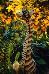 A wooden giraffe sculpture with intricate patterns stands among lush green foliage and white flowers. The background is a vibrant blend of golden bokeh, creating a warm and enchanting atmosphere.