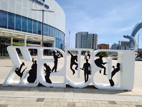 A large sculpture spelling out 'Fukuoka' in white letters with black silhouettes of musicians and dancers cut out from the letters. The structure is placed in an urban plaza with modern buildings in the background. A few people can be seen walking around, and a clear blue sky suggests a bright, sunny day.