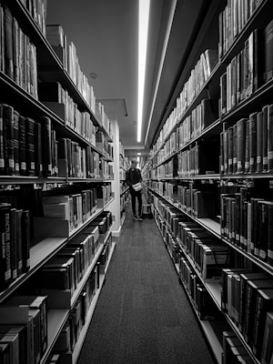A black and white image of a library aisle lined with tall bookshelves filled with books. A person stands in the middle, browsing a book, and the lighting above creates a dramatic, focused atmosphere.