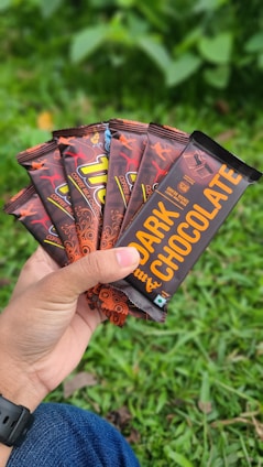 A hand is holding several chocolate bars with vibrant packaging that includes designs of dancing figures and bold text. The bars are labeled as 'dark chocolate' and 'coffee milk chocolate'. The background features green grass, suggesting an outdoor setting.