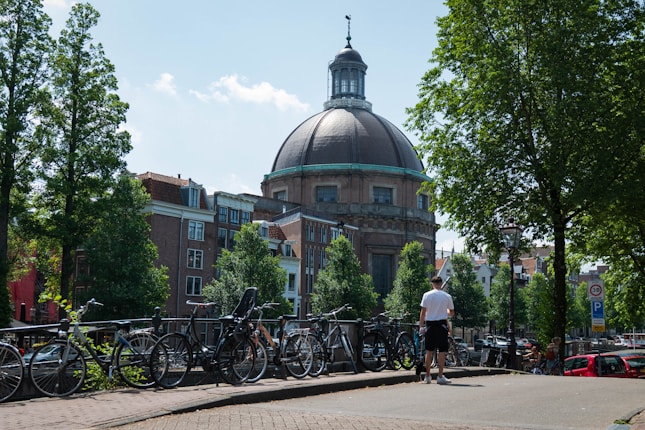 A large domed building in the background surrounded by trees and traditional brick buildings. In the foreground, several bicycles are parked along a canal railing, with a person walking near them. The scene is sunny with a clear blue sky.