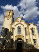 A historic church with ornate architecture, featuring arched windows and decorative moldings. The facade is lit by the sun, casting shadows from nearby trees. A clock tower is seen on the left with a cross on top, under a bright blue sky with scattered clouds.