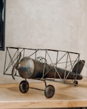 A vintage-style model biplane made of metal is resting on a wooden surface. The design features an old-fashioned propeller and spoked wheels, with an intricate frame structure. The background is a plain, textured wall.