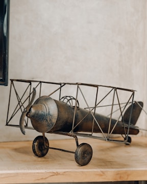 A vintage-style model biplane made of metal is resting on a wooden surface. The design features an old-fashioned propeller and spoked wheels, with an intricate frame structure. The background is a plain, textured wall.