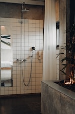 A modern bathroom featuring a white-tiled wall with a shower head and a handheld shower. A white, sheer curtain partially covers the area, and a plant is positioned on a concrete planter to the right. A glimpse into a bedroom with a white bed is visible through an open doorway, reflecting a minimalist design.