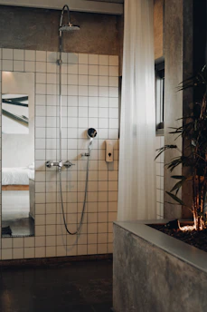 A modern bathroom featuring a white-tiled wall with a shower head and a handheld shower. A white, sheer curtain partially covers the area, and a plant is positioned on a concrete planter to the right. A glimpse into a bedroom with a white bed is visible through an open doorway, reflecting a minimalist design.