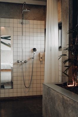 A modern bathroom featuring a white-tiled wall with a shower head and a handheld shower. A white, sheer curtain partially covers the area, and a plant is positioned on a concrete planter to the right. A glimpse into a bedroom with a white bed is visible through an open doorway, reflecting a minimalist design.