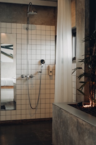 A modern bathroom featuring a white-tiled wall with a shower head and a handheld shower. A white, sheer curtain partially covers the area, and a plant is positioned on a concrete planter to the right. A glimpse into a bedroom with a white bed is visible through an open doorway, reflecting a minimalist design.