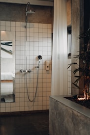 A modern bathroom featuring a white-tiled wall with a shower head and a handheld shower. A white, sheer curtain partially covers the area, and a plant is positioned on a concrete planter to the right. A glimpse into a bedroom with a white bed is visible through an open doorway, reflecting a minimalist design.