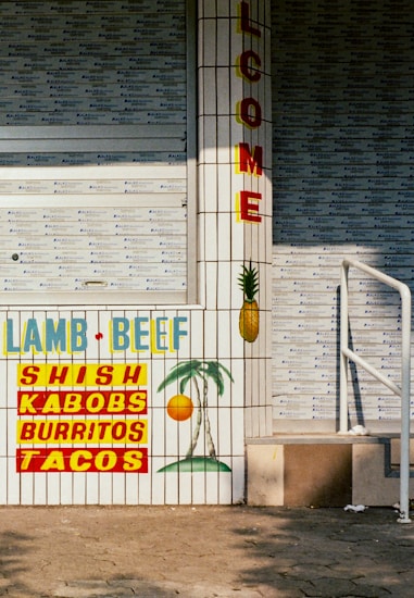 A tiled wall features colorful signs advertising various foods like lamb, beef, shish kabobs, burritos, and tacos. The word 'WELCOME' is prominently displayed vertically. A painted pineapple and a palm tree with a sun create a tropical theme.