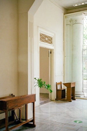 A spacious, sunlit interior featuring an open doorway leading into another room. The space has marble flooring and contains wooden furniture, including a desk and a simple chair. There is a green plant peeking from around the doorway and a small circular green sticker or decal on the floor.
