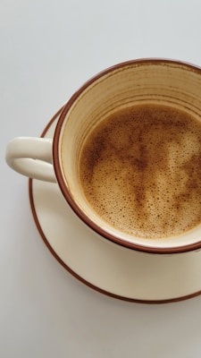 A ceramic mug filled with frothy coffee sits on a matching saucer. The mug features a light beige color with dark brown speckles and a simple, classic design.