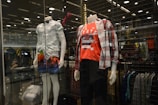 A clothing store display featuring two mannequins dressed in casual outfits. The left mannequin is wearing a short-sleeved leaf-patterned shirt paired with brightly colored shorts depicting a palm tree and sunset design. The right mannequin is dressed in a red graphic t-shirt with a plaid shirt worn open over it and dark pants. Behind them, shelves and racks of clothing are visible, and the display is enclosed in glass with lighting accents overhead.