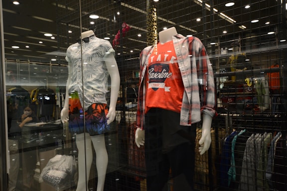 A clothing store display featuring two mannequins dressed in casual outfits. The left mannequin is wearing a short-sleeved leaf-patterned shirt paired with brightly colored shorts depicting a palm tree and sunset design. The right mannequin is dressed in a red graphic t-shirt with a plaid shirt worn open over it and dark pants. Behind them, shelves and racks of clothing are visible, and the display is enclosed in glass with lighting accents overhead.