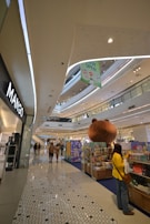 Inside a modern shopping mall, a large open space with multiple levels is visible. Shoppers walk along a tiled floor, passing various retail stores. A giant bear sculpture serves as a decorative centerpiece near a colorful display of merchandise. Bright, even lighting comes from overhead, reflecting off the smooth surfaces.