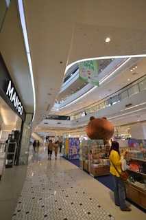 Inside a modern shopping mall, a large open space with multiple levels is visible. Shoppers walk along a tiled floor, passing various retail stores. A giant bear sculpture serves as a decorative centerpiece near a colorful display of merchandise. Bright, even lighting comes from overhead, reflecting off the smooth surfaces.