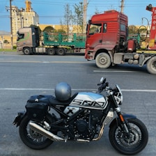A motorcycle with a helmet on the seat is parked on a road. In the background, there are two large trucks, one red and the other with green cargo frames, parked parallel to the street. Some trees and a building with industrial features are visible in the background under a blue sky.