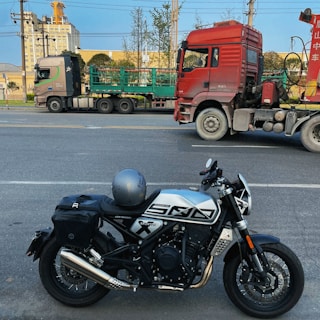 A motorcycle with a helmet on the seat is parked on a road. In the background, there are two large trucks, one red and the other with green cargo frames, parked parallel to the street. Some trees and a building with industrial features are visible in the background under a blue sky.
