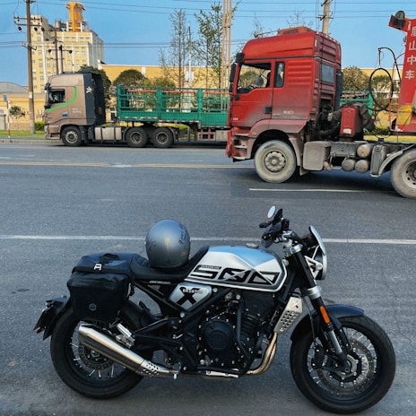 A motorcycle with a helmet on the seat is parked on a road. In the background, there are two large trucks, one red and the other with green cargo frames, parked parallel to the street. Some trees and a building with industrial features are visible in the background under a blue sky.