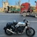 A motorcycle with a helmet on the seat is parked on a road. In the background, there are two large trucks, one red and the other with green cargo frames, parked parallel to the street. Some trees and a building with industrial features are visible in the background under a blue sky.