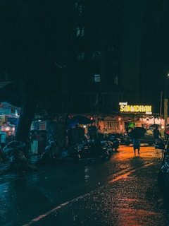 A dimly lit urban street scene at night with wet reflective pavement. There is a neon sign that reads 'Samadhan' in the background, with some people carrying umbrellas walking down the street. The area appears bustling with parked scooters and small street-side shops.