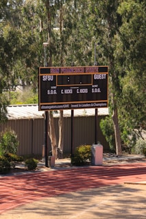 An outdoor scoreboard for San Francisco State is seen among trees and greenery. The board displays sections for shots on goal and corner kicks, with areas for both home and guest teams. It is situated near a running track with a building in the background.