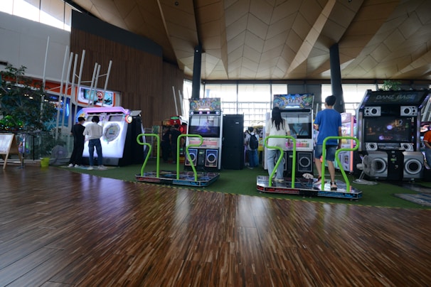 A vibrant arcade setting with several gaming machines lined up, including dance and rhythm game setups. People are engaged with the games, absorbed in their activities. The flooring is wooden, and the ceiling has a geometric design. Natural light filters through large windows, creating a lively atmosphere.