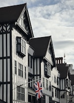 A traditional building with half-timbered architecture featuring dark wooden beams and white plaster, adorned with an old-fashioned window design. A British flag hangs prominently from the building's fa&ccedil;ade. The sky is partly cloudy, and there is a decorative weather vane or ornament on the roof.