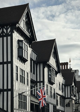 A traditional building with half-timbered architecture featuring dark wooden beams and white plaster, adorned with an old-fashioned window design. A British flag hangs prominently from the building's fa&ccedil;ade. The sky is partly cloudy, and there is a decorative weather vane or ornament on the roof.