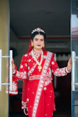 A woman gracefully wearing a bright red saree with gold embroidery, standing near a temple entrance.