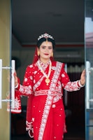 A woman dressed in traditional attire stands at a doorway, wearing a vibrant red saree adorned with intricate white embroidery. Her jewelry includes gold earrings, a necklace, and a decorative headpiece. Her hands show detailed henna designs, and she is smiling confidently.