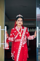 A woman dressed in traditional attire stands at a doorway, wearing a vibrant red saree adorned with intricate white embroidery. Her jewelry includes gold earrings, a necklace, and a decorative headpiece. Her hands show detailed henna designs, and she is smiling confidently.