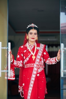 A woman dressed in traditional attire stands at a doorway, wearing a vibrant red saree adorned with intricate white embroidery. Her jewelry includes gold earrings, a necklace, and a decorative headpiece. Her hands show detailed henna designs, and she is smiling confidently.