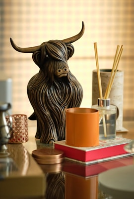 A detailed wooden sculpture of a bull with long, flowing hair serves as the centerpiece. Surrounding it are various modern decor items including a reed diffuser, a candle, and a reflective glass surface enhancing the scene. The lighting is warm, with a soft, blurred grid pattern in the background creating an elegant ambiance.