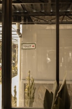 A view of a toilet sign with an arrow on a wall, indicating the direction to the restroom. There are indoor plants visible at the bottom, and the scene is viewed through a window, adding reflections and a layer of complexity to the image. The setting appears to be in a functional, perhaps industrial location with pipes and metal structures in the background.